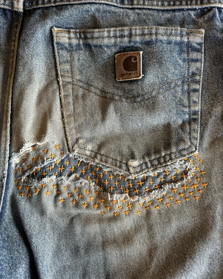 Carhartt jeans back pocket with a long tear backed by a denim patch, held with rows of orange cross-stitch.