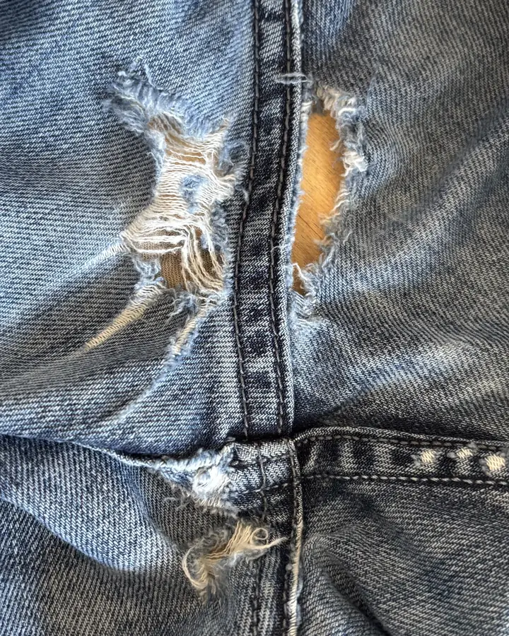 Light-wash jeans worn into open holes on both sides of the seam junction, with loose threads across the gaps.