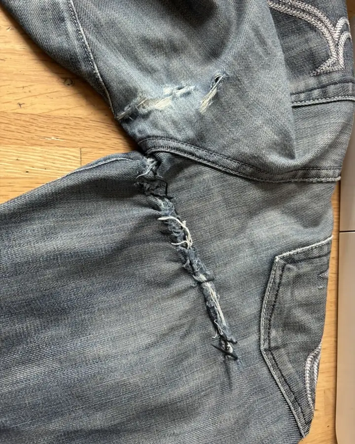 Pale jeans on a wooden floor with a long frayed split running below the seam and a smaller tear above it.