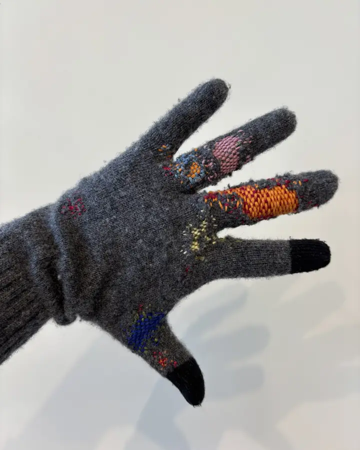 The same grey wool glove from the side, showing orange, pink and blue darns along the fingers and thumb.