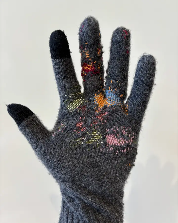 Grey wool glove seen palm-on, with patches of multicoloured darning across the palm and fingers.