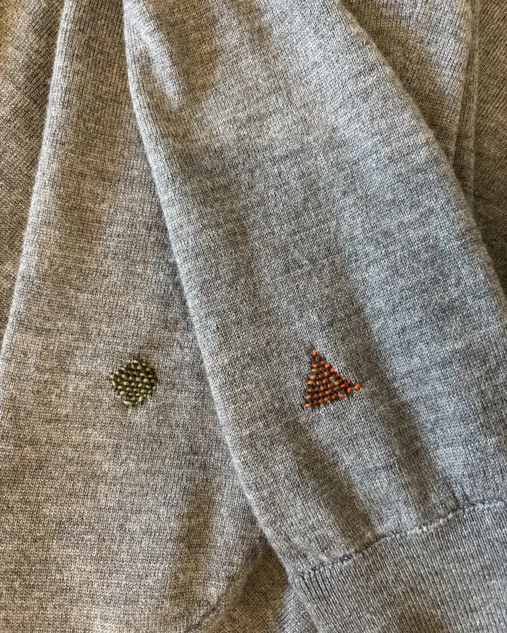 Grey wool sweater sleeves with a small green circular darn and a small orange triangular darn.
