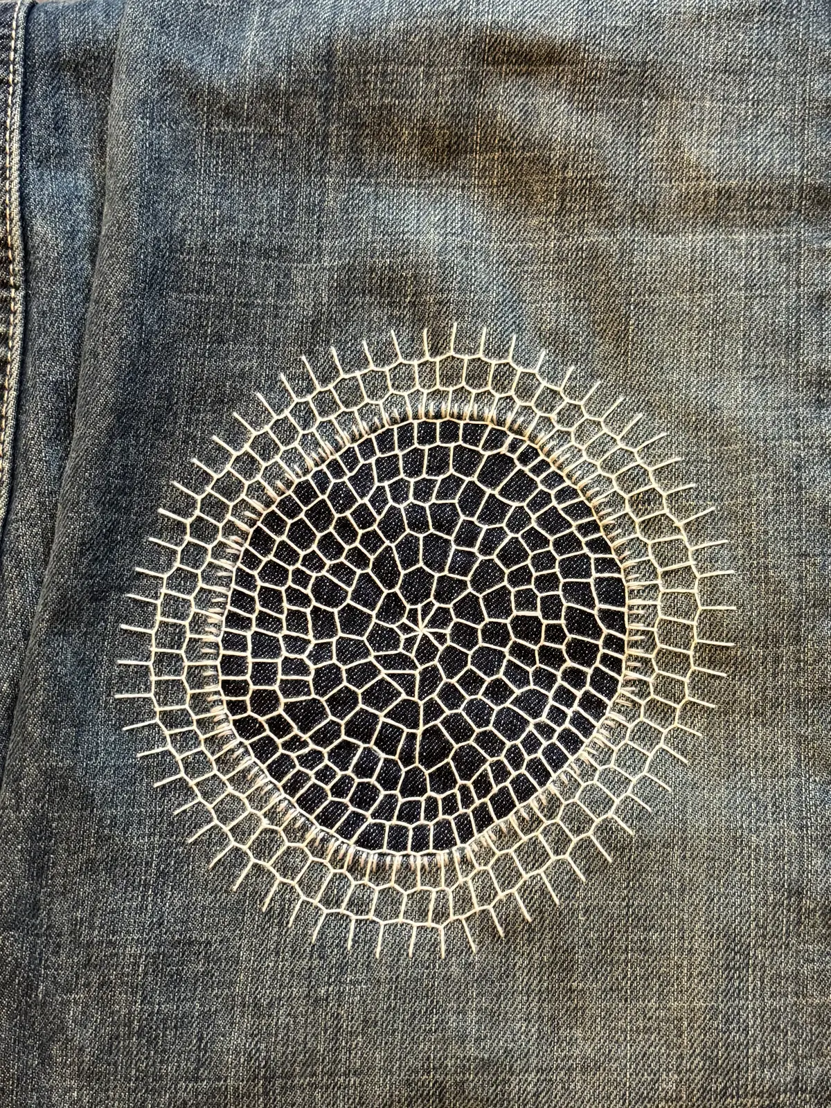 A worn patch of denim covered by a large sunburst darn worked in cream thread.