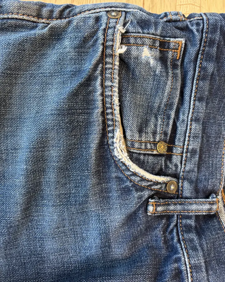 Front jeans pocket with the curved opening frayed to a pale white edge and beginning to split.
