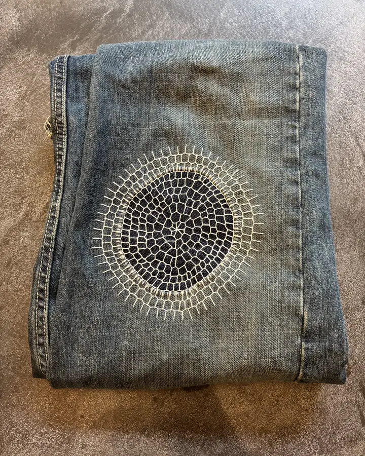 Folded jeans with a large circular sunburst darn in cream thread covering a worn patch of denim.